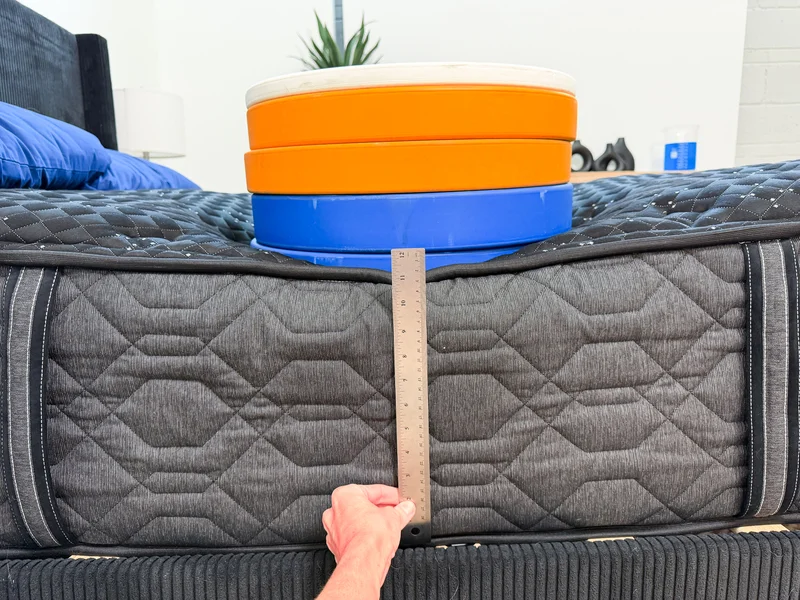 A stack of weight plates are sitting a top the Beautyrest Black and a tester is holding up a ruler to evaluate weighted edge support.